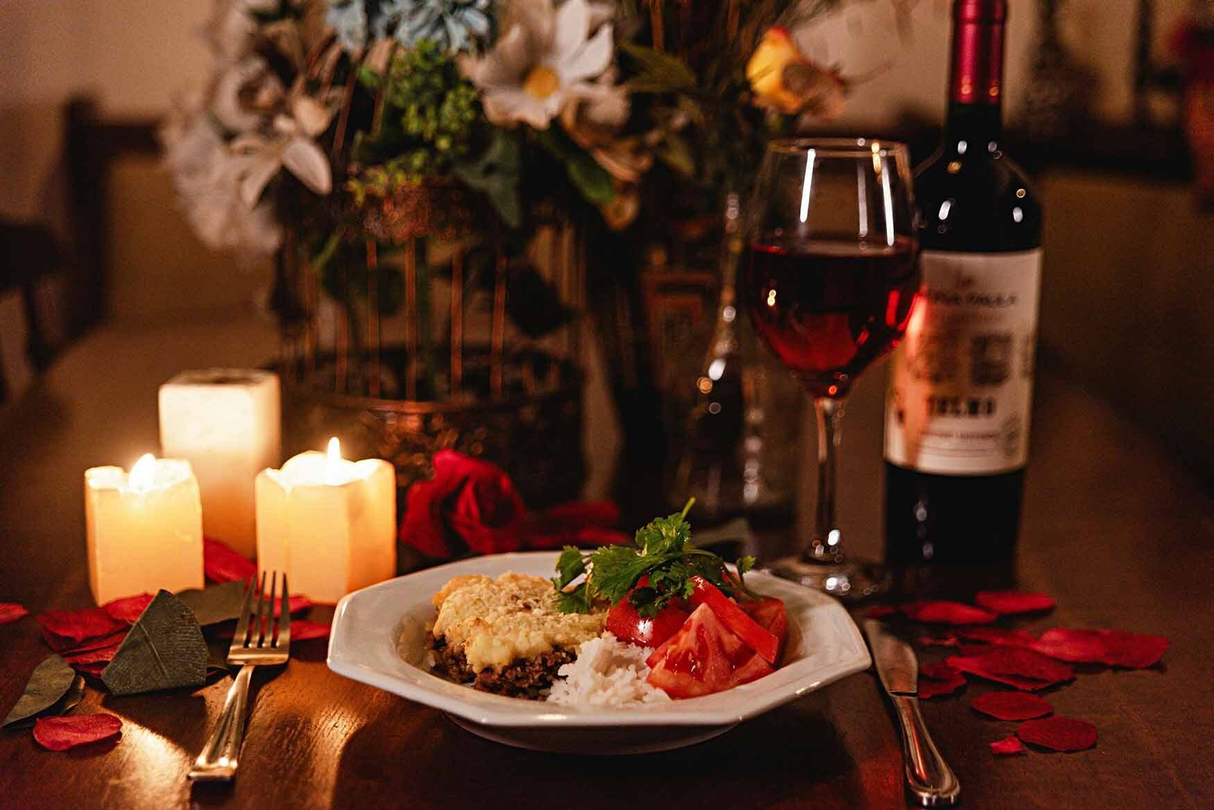 Cena romántica con platillo, velas, copa y botella de vino sobre mesa de madera con pétalos de rosa.